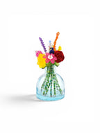 A small, clear light blue glass vase containing a bouquet of colorful beaded flowers. The stems are made of green beads, while the blossoms feature various shapes and vibrant colors including red, yellow, pink, purple, and orange.