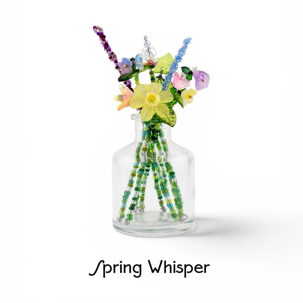 A clear glass bottle containing a bouquet of handcrafted beaded flowers in yellow, purple, pink, and blue, with green beaded stems. The text Spring Whisper is printed below the bottle. The arrangement features various glass beads.