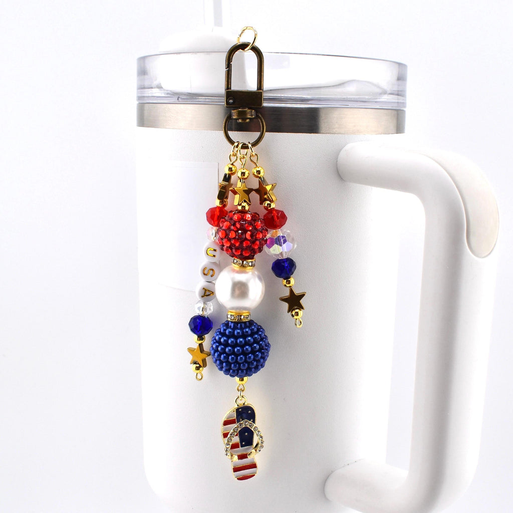 A patriotic keychain charm with red, white, and blue beads, gold stars, and a flip-flop charm featuring the American flag. The charm is attached to a bronze-colored clasp and is hanging on a white mug.