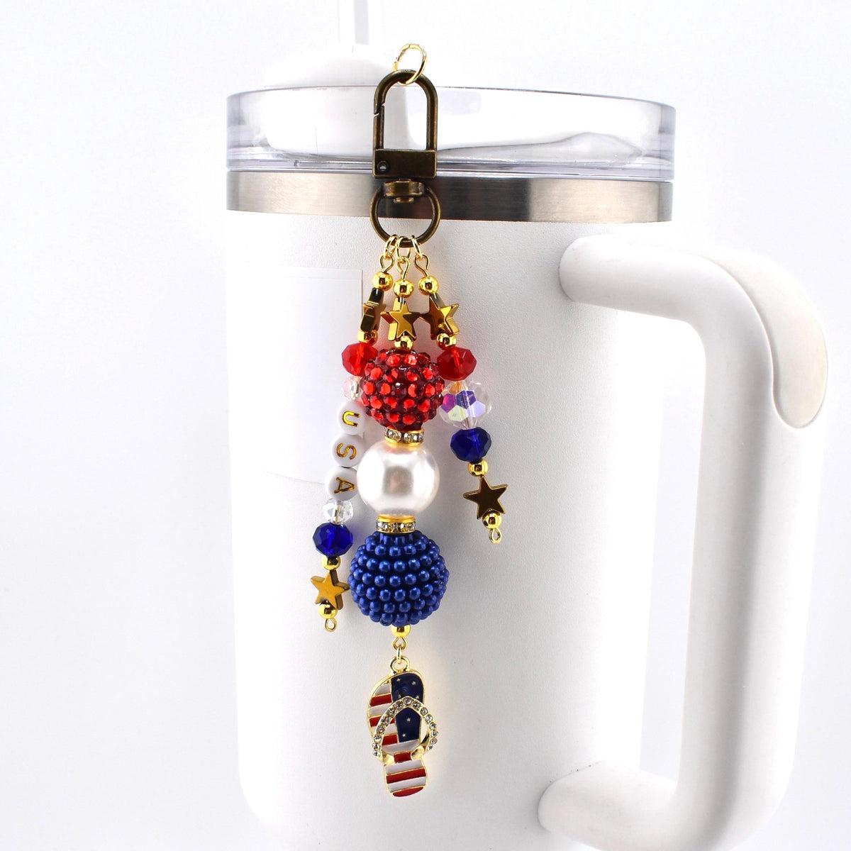 A patriotic keychain charm with red, white, and blue beads, gold stars, and a flip-flop charm featuring the American flag. The charm is attached to a bronze-colored clasp and is hanging on a white mug.
