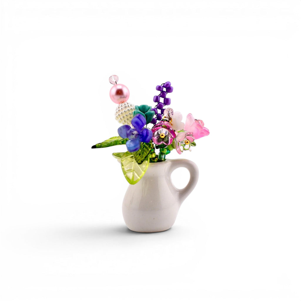 A miniature white ceramic pitcher filled with a decorative arrangement of colorful glass and plastic beads shaped like flowers and leaves. The bouquet includes pink, purple, blue, and green elements with a prominent pearl-like bead.