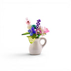 A miniature white ceramic pitcher filled with a decorative arrangement of colorful glass and plastic beads shaped like flowers and leaves. The bouquet includes pink, purple, blue, and green elements with a prominent pearl-like bead.