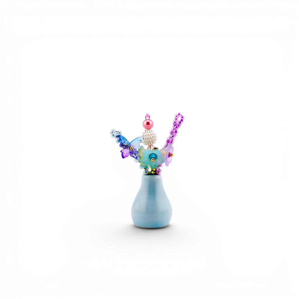 A small, light blue ceramic vase containing a decorative arrangement of colorful beads and butterfly-shaped accents. The arrangement features a pink pearl, a white textured sphere, and purple, blue, and green translucent elements.
