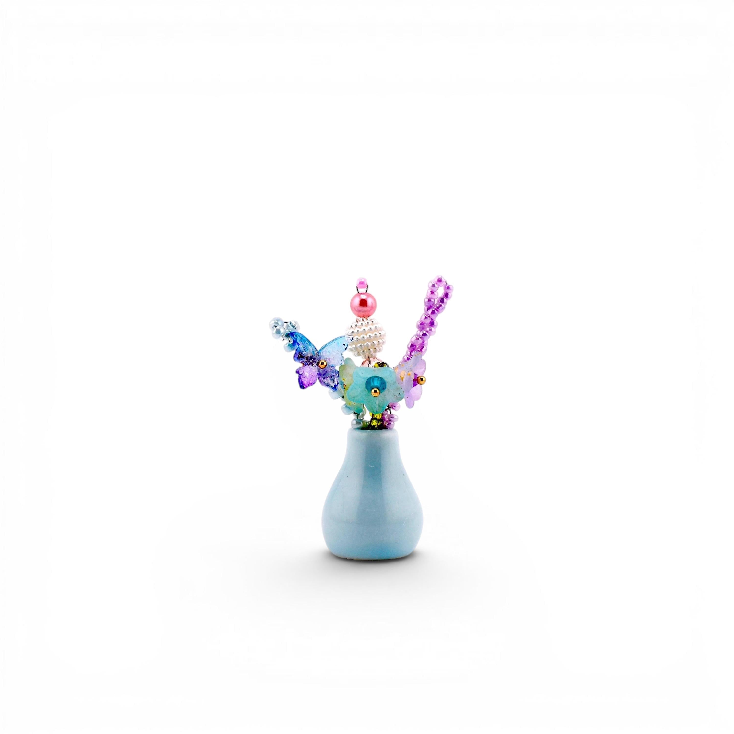 A small, light blue ceramic vase containing a decorative arrangement of colorful beads and butterfly-shaped accents. The arrangement features a pink pearl, a white textured sphere, and purple, blue, and green translucent elements.