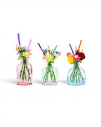 Three small glass bottles in pink, clear, and light blue, each containing a bouquet of handcrafted beaded flowers. The stems are made of green beads, and the blossoms feature various colors including yellow, red, purple, and pink.