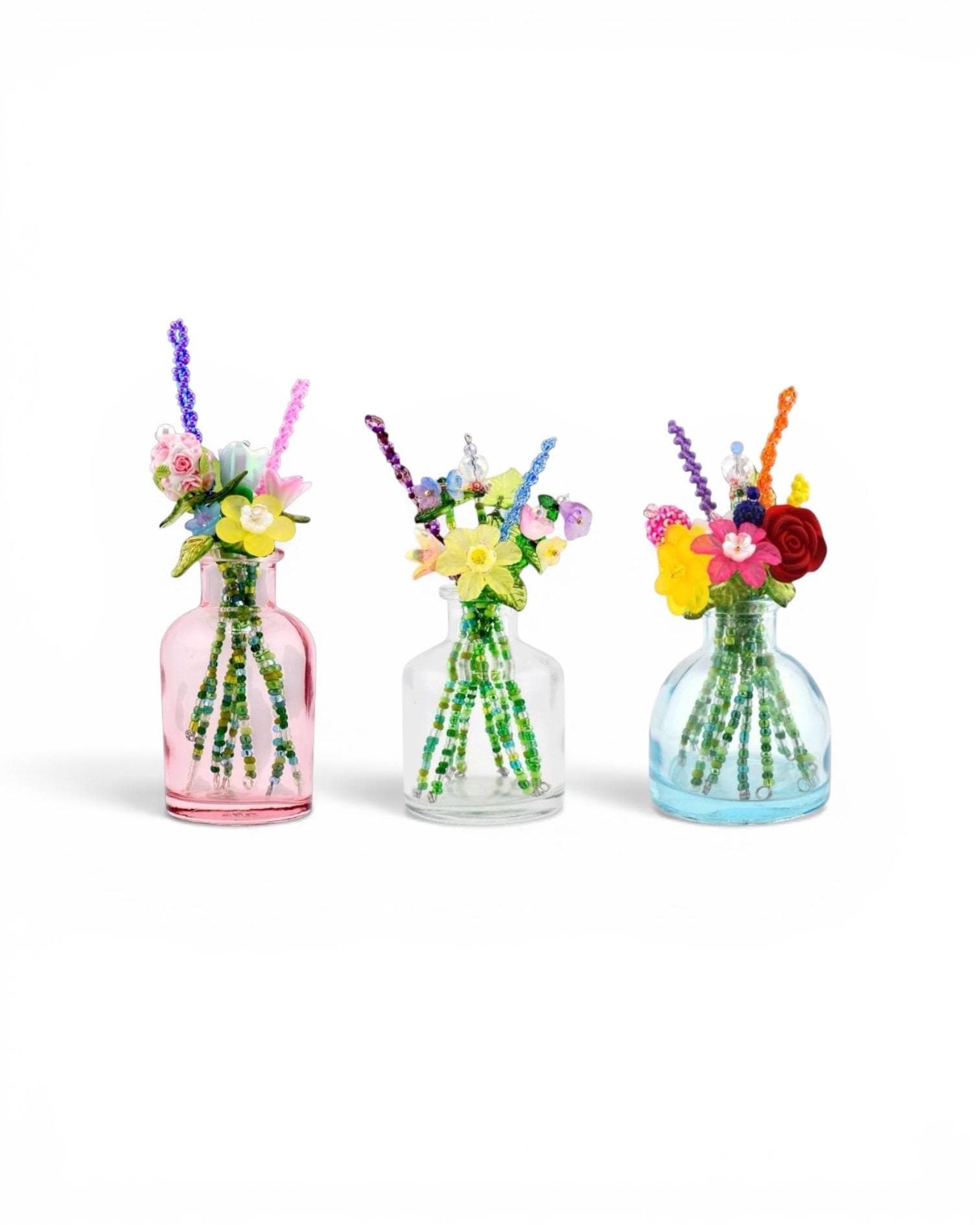 Three small glass bottles in pink, clear, and light blue, each containing a bouquet of handcrafted beaded flowers. The stems are made of green beads, and the blossoms feature various colors including yellow, red, purple, and pink.