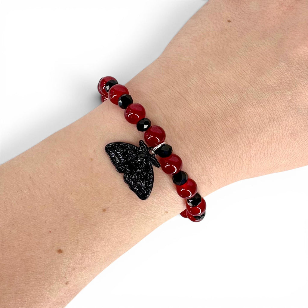 A beaded bracelet featuring alternating smooth red spherical beads and smaller faceted black beads. A black moth-shaped charm hangs from the center of the bracelet. The beads are strung on an elastic cord, creating a circular design.