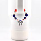 A beaded bracelet featuring red, white, and blue beads, star-shaped beads with American flag patterns, and a silver-toned bow charm. The bracelet is constructed with alternating faceted, smooth, and rhinestone-encrusted spacer beads.
