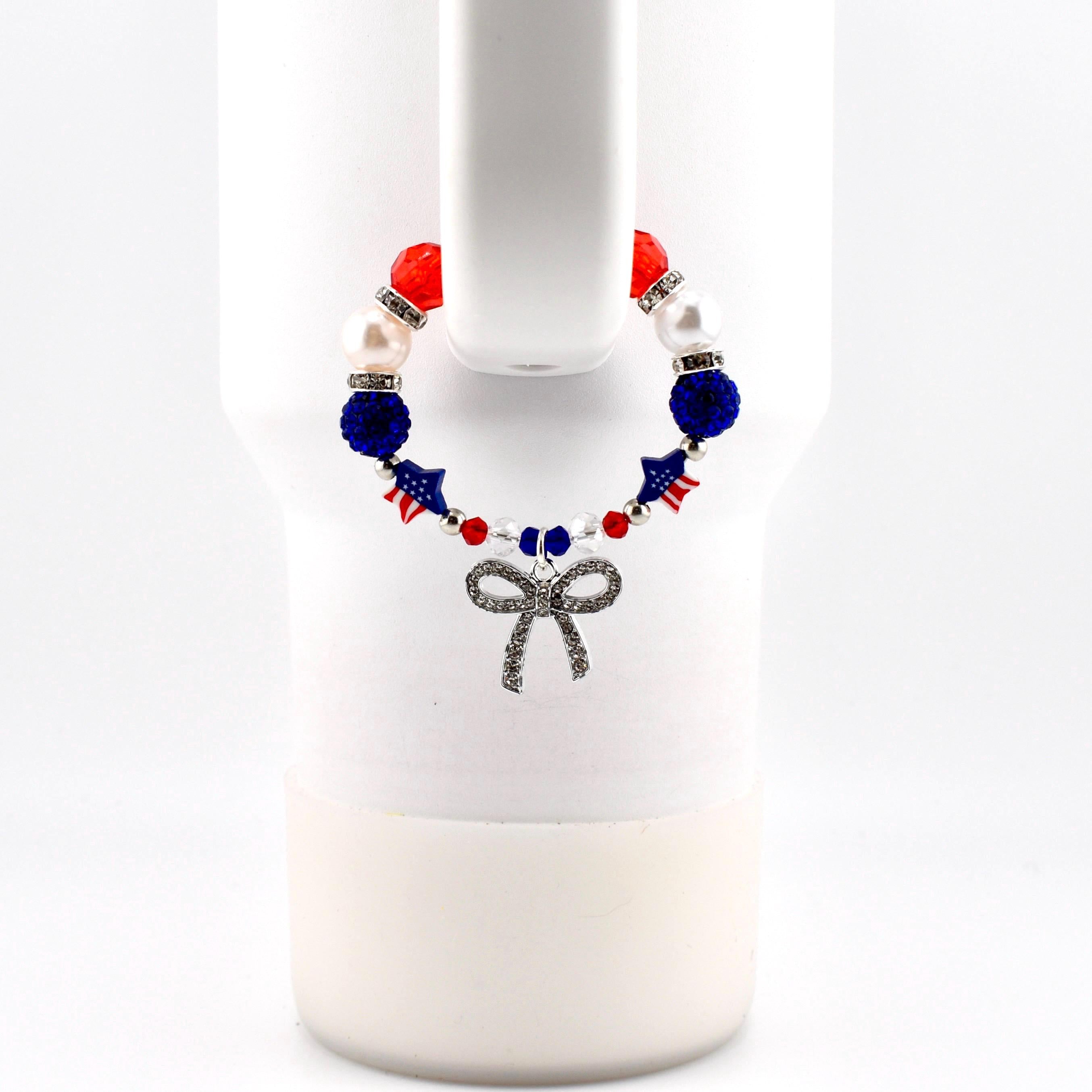 A beaded bracelet featuring red, white, and blue beads, star-shaped beads with American flag patterns, and a silver-toned bow charm. The bracelet is constructed with alternating faceted, smooth, and rhinestone-encrusted spacer beads.