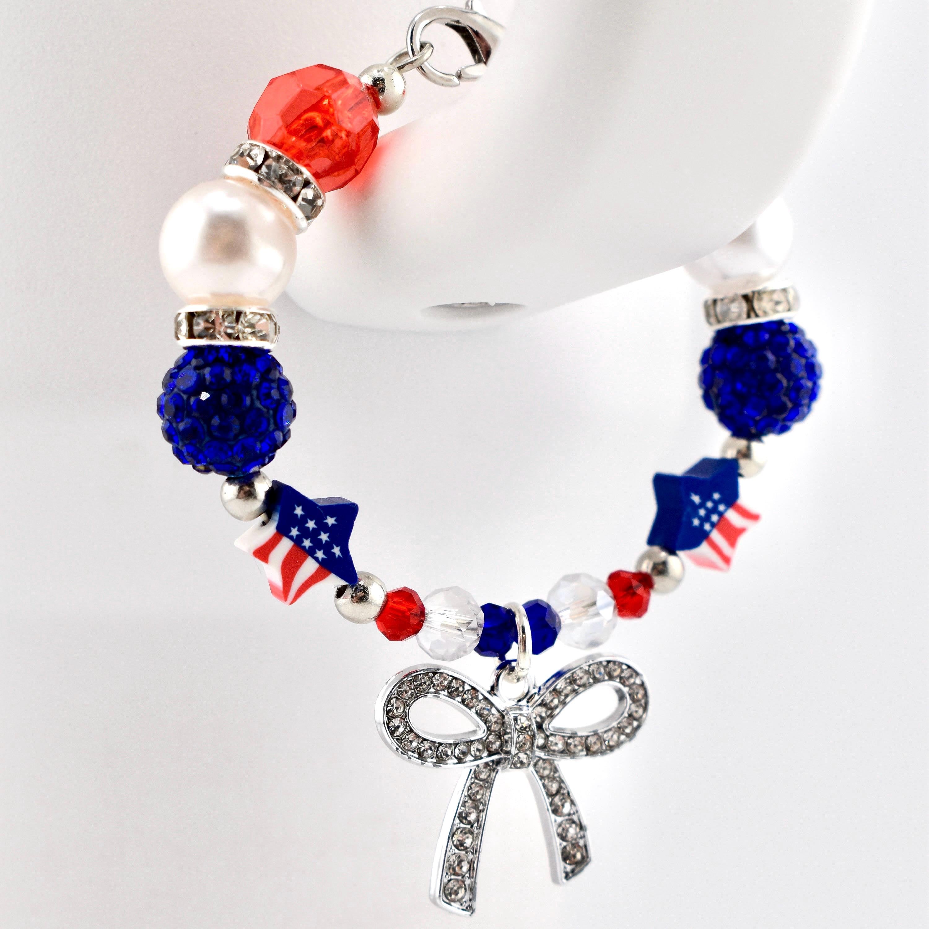 A beaded tumbler handle bracelet featuring red, white, and blue beads, including faceted red beads, white pearls, blue glitter spheres, and star-shaped beads with American flag patterns. A silver-toned bow charm encrusted with clear rhinestones hangs.