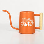 An orange metal watering can with a long, slender spout and a light-colored wooden handle. The side of the watering can features a white graphic of various wildflowers and the name Jacki written in a cursive script font.