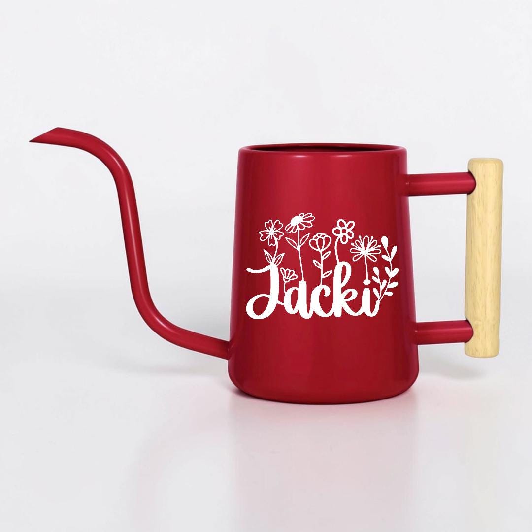 A red metal watering can with a long, slender spout and a light-colored wooden handle. The side of the watering can features a white graphic of various wildflowers and the name Jacki written in a cursive script font.