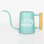 A light blue metal watering can with a long, thin spout and a light-colored wooden handle. The side features a white graphic of four potted plants above the text Kamilah's GREENHOUSE in a mix of script and block lettering.