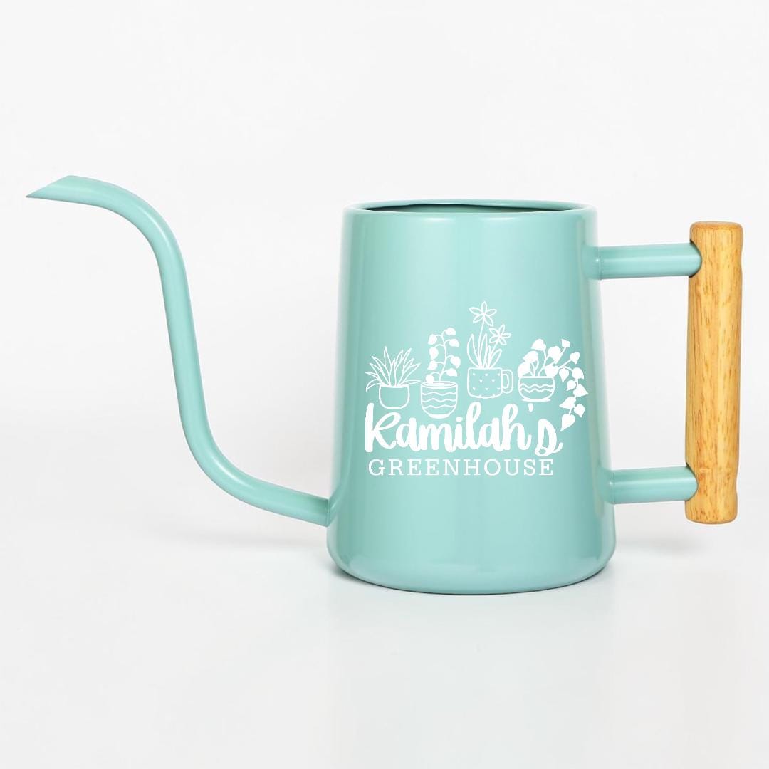 A light blue metal watering can with a long, thin spout and a light-colored wooden handle. The side features a white graphic of four potted plants above the text Kamilah's GREENHOUSE in a mix of script and block lettering.