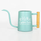 A light blue metal watering can with a long, thin spout and a light wood-toned handle. The side features a white line art graphic of five potted plants above the white text Custom Text and OPTIONAL LINE 2 in a script font.
