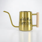 A gold-colored stainless steel watering can with a long, slender spout and a light-colored wooden handle. The body features a black line art graphic of five potted plants with the text Custom Text and OPTIONAL LINE 2 printed below.