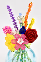 A decorative bouquet of artificial flowers and stems crafted from colorful beads, crystals, and plastic floral elements. The arrangement features a red rose, yellow and pink blossoms, and tall beaded stems in purple and orange.
