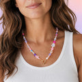 A colorful beaded necklace with a gold clasp. The necklace features a variety of beads in shades of pink, purple, blue, and white. The beads are arranged in a pattern with flower-shaped accents.