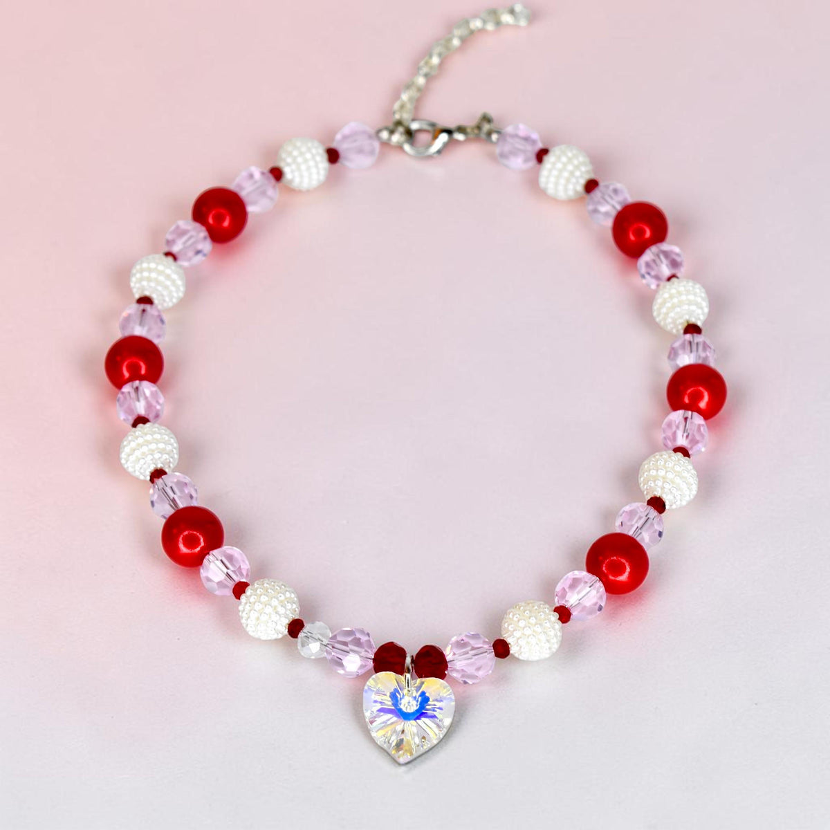 A beaded necklace featuring a repeating pattern of opaque white spheres, translucent light pink faceted beads, and solid red spheres. A clear, iridescent, heart-shaped pendant hangs from the center, attached by a silver-tone metal chain.