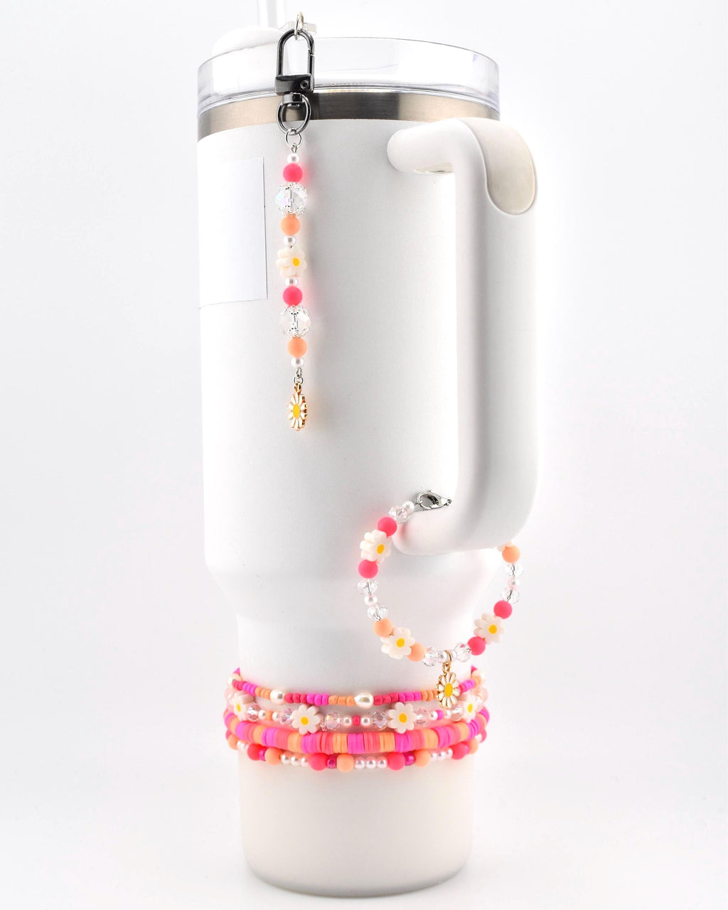 A white insulated tumbler with a handle, adorned with a beaded charm featuring pink, orange, and clear beads with a small daisy pendant, a matching beaded bracelet, and three stacked beaded bracelets in shades of pink and orange.