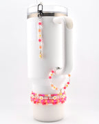A white insulated tumbler with a handle, adorned with a beaded charm featuring pink, orange, and clear beads with a small daisy pendant, a matching beaded bracelet, and three stacked beaded bracelets in shades of pink and orange.