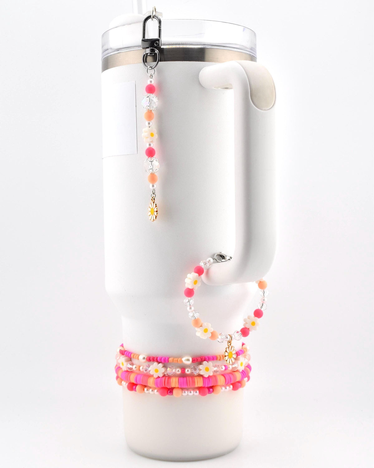 A white insulated tumbler with a handle, adorned with a beaded charm featuring pink, orange, and clear beads with a small daisy pendant, a matching beaded bracelet, and three stacked beaded bracelets in shades of pink and orange.