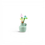 A small, light green ceramic vase with a single side handle, containing a decorative arrangement of multi-colored glass and crystal flowers in shades of pink, blue, yellow, and green, featuring various bead shapes and wire stems.