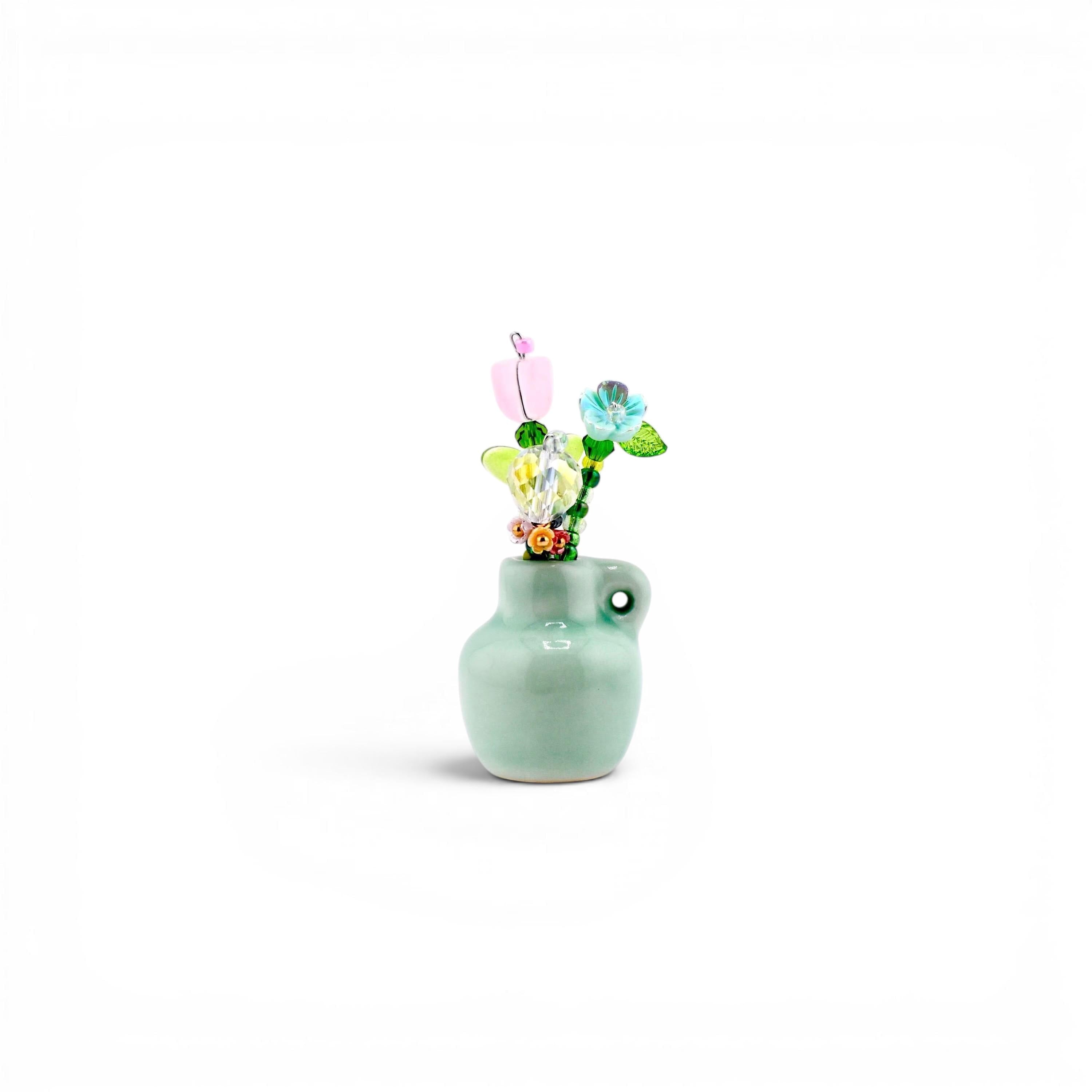 A small, light green ceramic vase with a single side handle, containing a decorative arrangement of multi-colored glass and crystal flowers in shades of pink, blue, yellow, and green, featuring various bead shapes and wire stems.