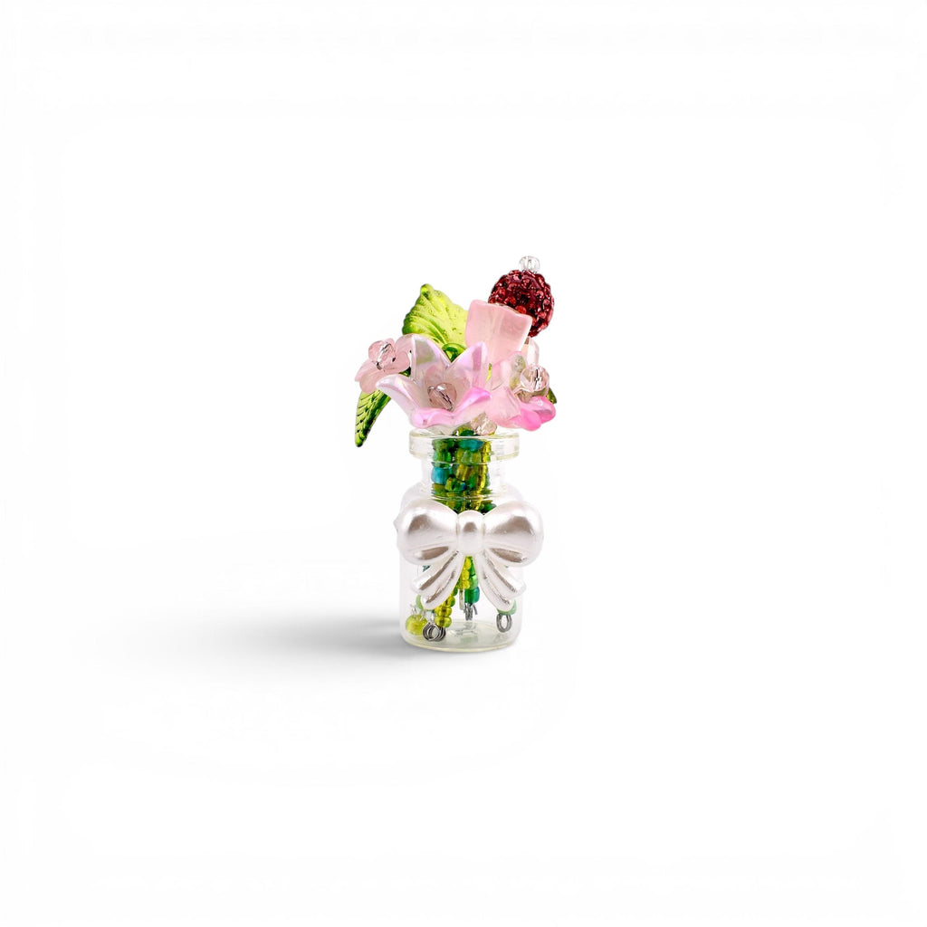 A miniature clear glass bottle containing a small bouquet of artificial flowers, including pink blossoms, green leaves, and a dark red textured sphere. The bottle is decorated with a white plastic bow attached to the front neck.