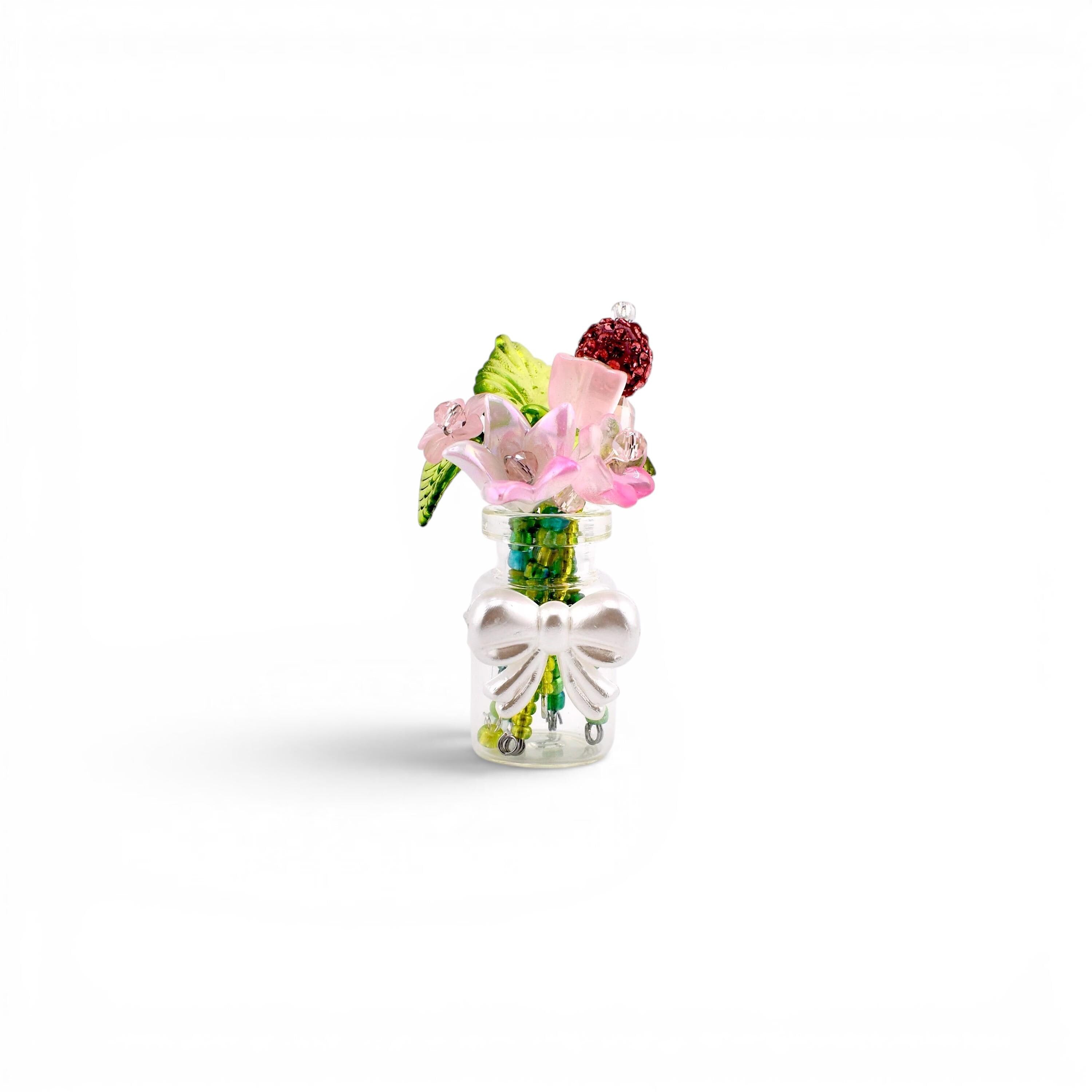 A miniature clear glass bottle containing a small bouquet of artificial flowers, including pink blossoms, green leaves, and a dark red textured sphere. The bottle is decorated with a white plastic bow attached to the front neck.
