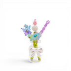 A small clear glass bottle decorated with a white bow, containing several decorative sticks topped with colorful beads, a blue butterfly, and light blue flower-shaped elements in shades of pink, purple, blue, and green.