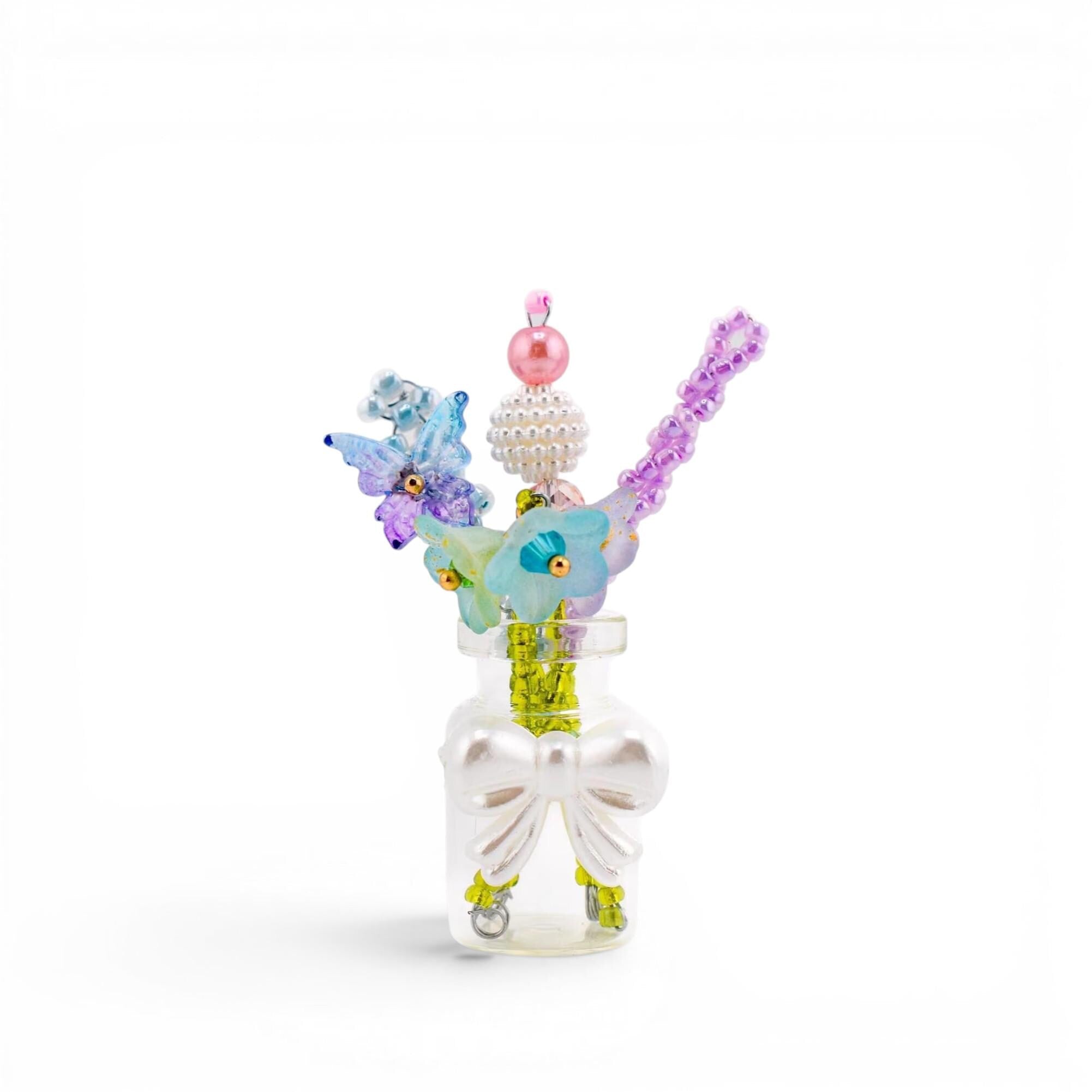 A small clear glass bottle decorated with a white bow, containing several decorative sticks topped with colorful beads, a blue butterfly, and light blue flower-shaped elements in shades of pink, purple, blue, and green.
