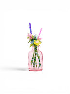 A small, translucent pink glass bottle containing a bouquet of artificial flowers made from colorful beads and wire. The arrangement features yellow, pink, and blue floral shapes with green beaded stems extending into the bottle.