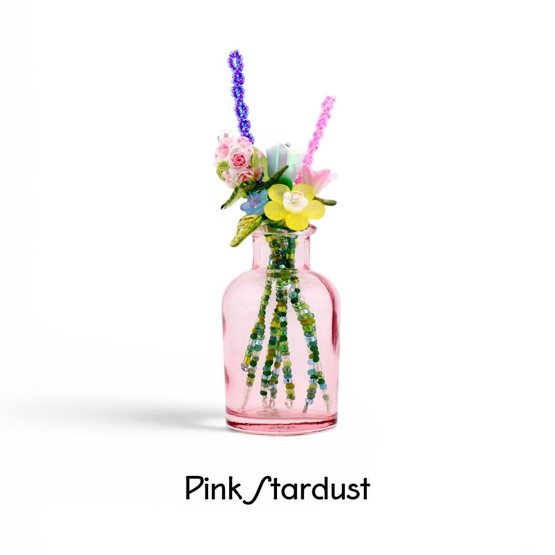 A small pink glass bottle containing a bouquet of artificial flowers made from colorful beads and plastic petals. The stems are crafted from green beads. The text Pink Stardust is printed in black below the bottle on a white background.