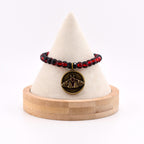 A beaded bracelet featuring translucent red and black spherical beads. A circular antique brass-toned pendant hangs from the center, displaying an embossed moth design with crescent moon motifs and a small red stone at the top.