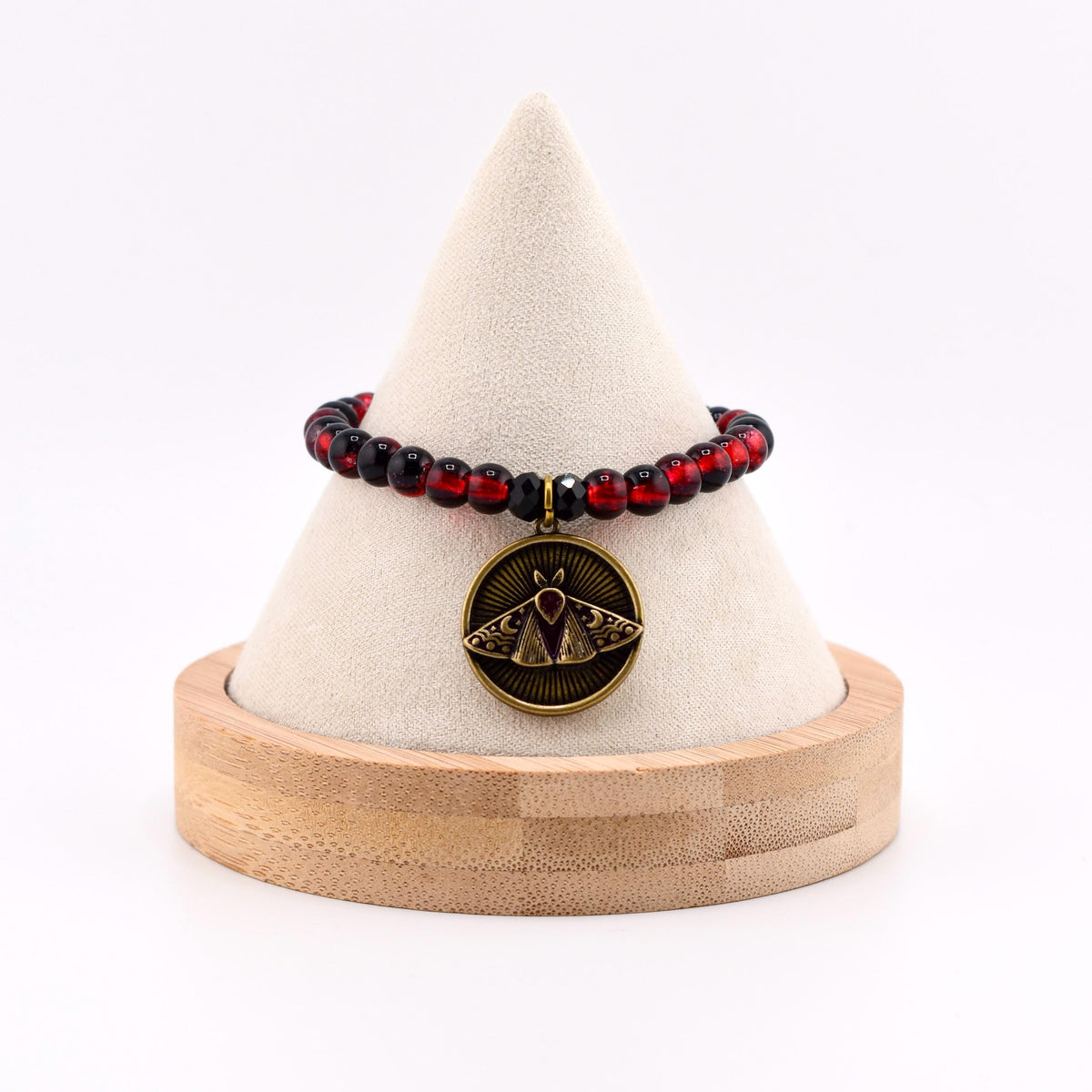 A beaded bracelet featuring translucent red and black spherical beads. A circular antique brass-toned pendant hangs from the center, displaying an embossed moth design with crescent moon motifs and a small red stone at the top.