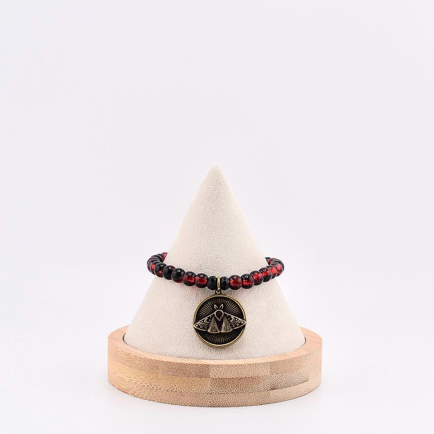 A beaded bracelet featuring alternating translucent red and opaque black spherical beads. A circular bronze-toned metal pendant hangs from the center, displaying an embossed moth design with small crescent moon details on its wings.