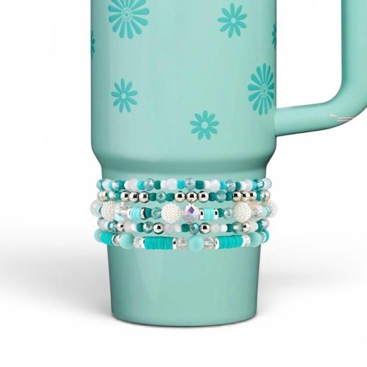 A light teal tumbler featuring a repeating pattern of small dark teal daisies. Wrapped around the lower section is a stack of five beaded bracelets in shades of turquoise, white, and silver, including round, faceted, and flat disc beads.