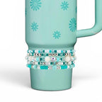 A light teal tumbler featuring a repeating pattern of small dark teal daisies. Wrapped around the lower section is a stack of five beaded bracelets in shades of turquoise, white, and silver, including round, faceted, and flat disc beads.