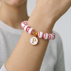 A beaded bracelet featuring white spherical beads with red stitching patterns resembling a baseball. The bracelet includes a gold-toned circular charm with a white center and the capital letter P in gold, accented by two gold-toned beads.