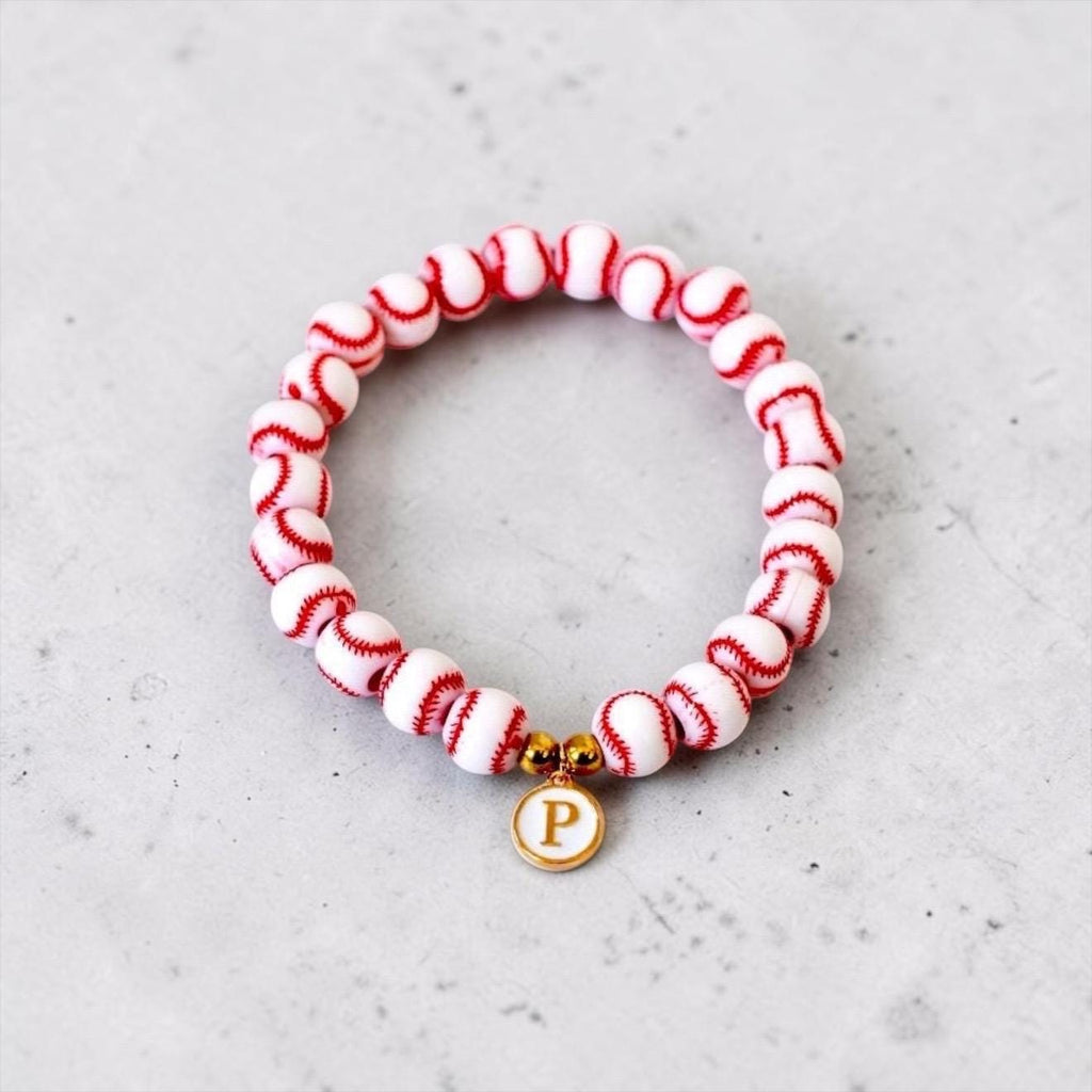 A circular stretch bracelet composed of white spherical beads with red stitched patterns resembling baseballs. The bracelet features two small gold-toned metallic spacer beads and a round white charm with a gold-toned letter P.