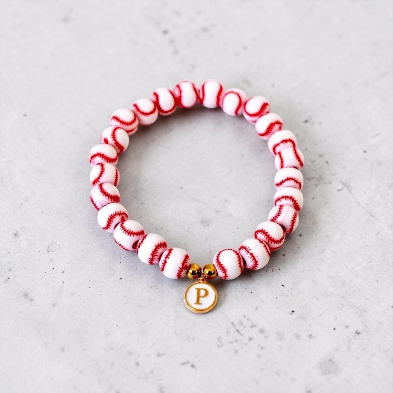 A circular stretch bracelet composed of white spherical beads with red stitched patterns resembling baseballs. The bracelet features two small gold-toned metallic spacer beads and a round white charm with a gold-toned letter P.