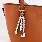 A bag charm featuring a silver-toned metal lobster clasp and ring. Three dangling strands consist of pink and light blue beads, clear crystal-like spacers, a small white seashell, and a white unicorn charm. One strand features white cube beads with black letters spelling BEACH BABE.