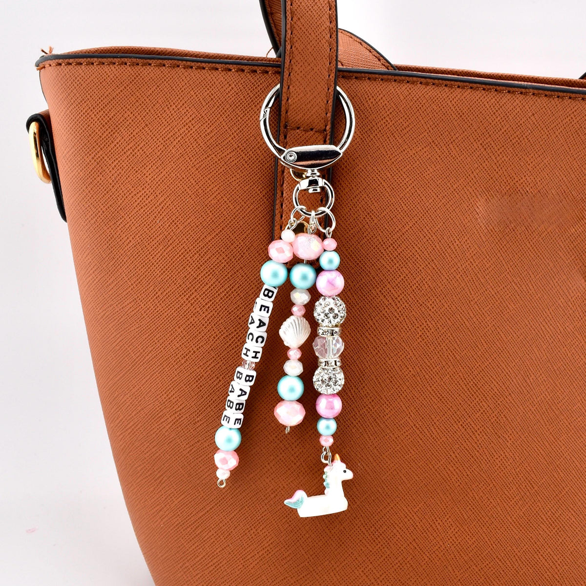 A bag charm featuring a silver-toned metal lobster clasp and ring. Three dangling strands consist of pink and light blue beads, clear crystal-like spacers, a small white seashell, and a white unicorn charm. One strand features white cube beads with black letters spelling BEACH BABE.