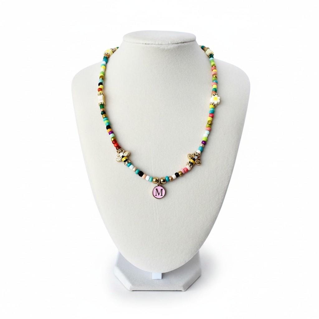 A beaded necklace featuring a variety of small, multicolored glass beads, two gold-toned bee charms, and a central circular pendant with a pink background and the letter M in a dark font, all strung on a flexible cord.