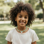 May include: A young person wearing a beaded necklace with green, yellow, and clear beads. The necklace has a heart-shaped pendant. The person is wearing a light-colored t-shirt and smiling. The background is a blurred outdoor scene.