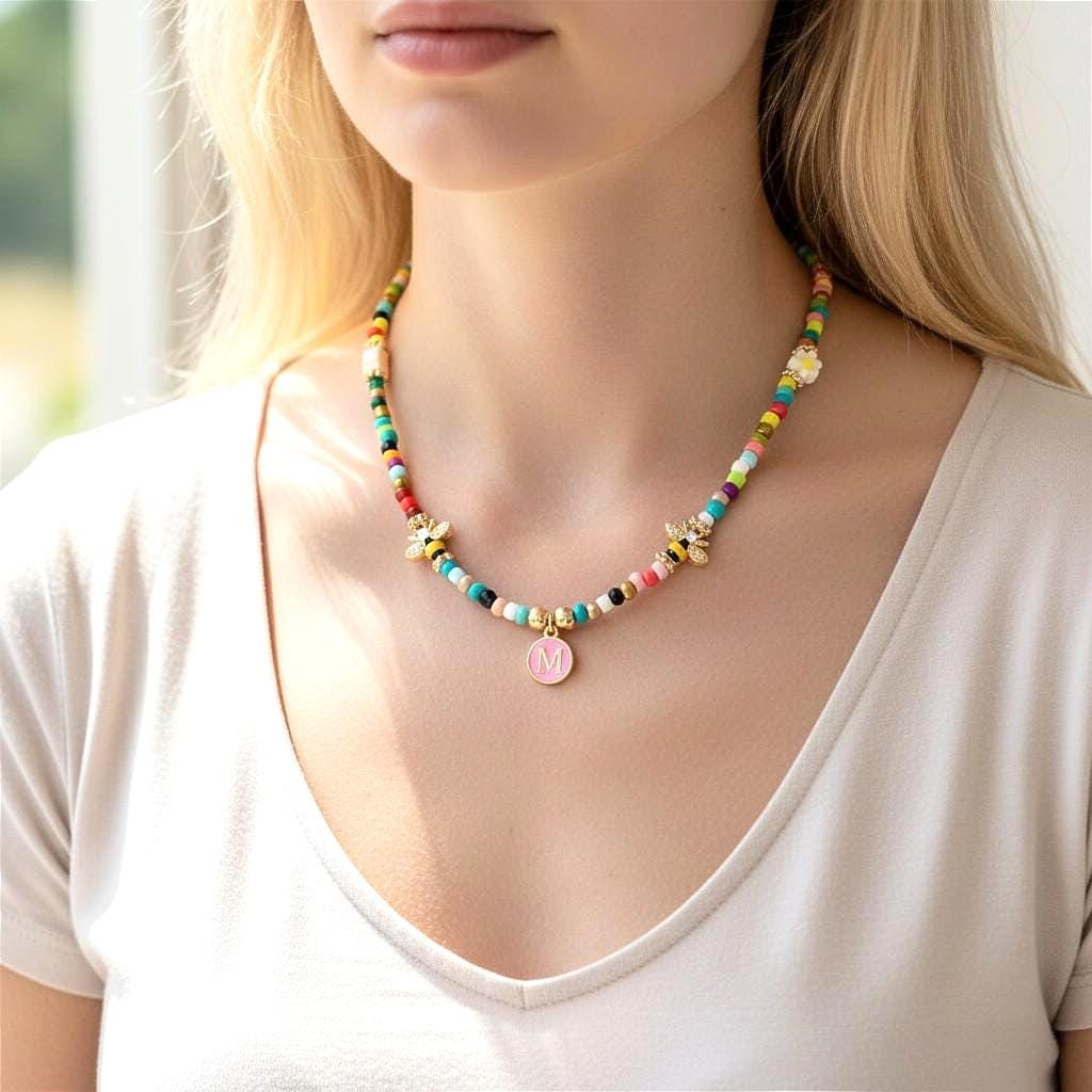 A beaded necklace featuring a variety of colorful small beads, two gold-toned bee charms, and a central round pendant with a light pink enamel finish and the letter M in the center. The necklace is worn around the neck.