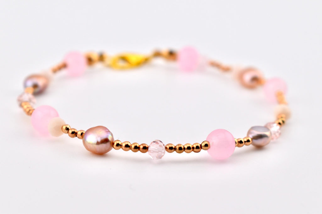 A beaded bracelet featuring a mix of round pink beads, iridescent pearls, small faceted crystals, and tiny metallic gold-tone spacer beads. The bracelet is secured with a gold-tone lobster claw clasp, creating a delicate and layered design.