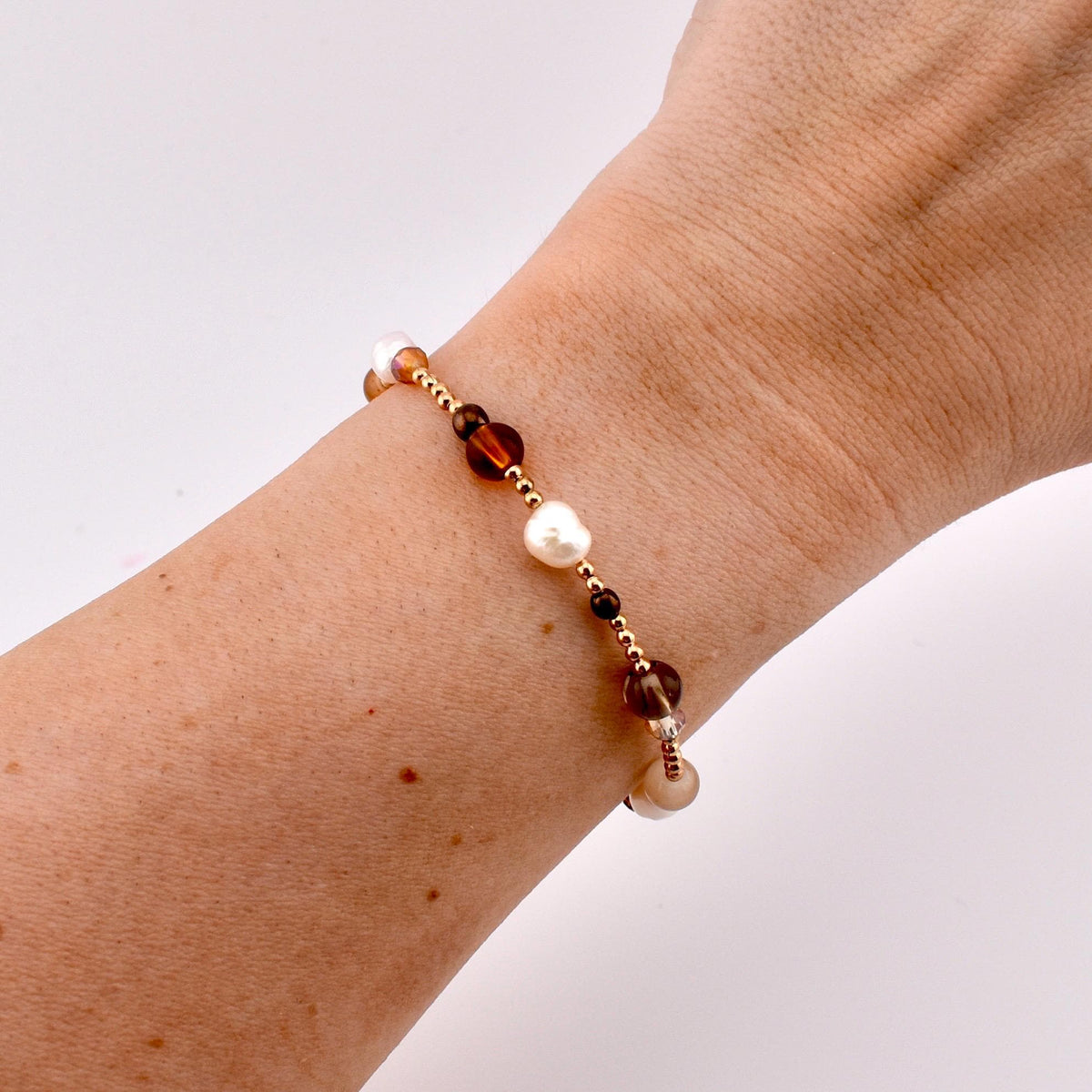 A beaded bracelet featuring a mix of small gold-toned metallic beads, white irregular freshwater pearls, and translucent brown glass beads. The beads are arranged in a repeating pattern along a thin, flexible cord worn around the wrist.