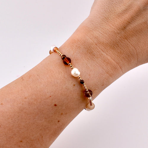 A beaded bracelet featuring a mix of small gold-toned metallic beads, white irregular freshwater pearls, and translucent brown glass beads. The beads are arranged in a repeating pattern along a thin, flexible cord worn around the wrist.